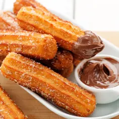 Churros posypane cukrem, jeden zanurzony w czekoladzie, obok miseczki z kremem czekoladowym.